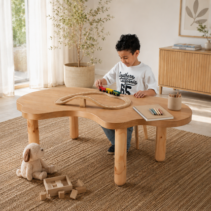 A child sits at a curved, low wooden table in a bright, modern room, playing with a wooden train track and toy train. The table has a natural light wood finish and is styled with a plush rabbit toy and wooden blocks on a woven rug.
